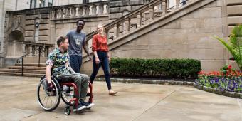 three people, one in a wheelchair and two walking, outside of the cathedral of learning