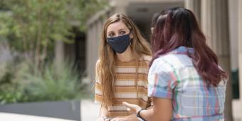 two woman, one facing the camera with a mask on, and one with her back to the camera, are talking outside